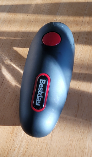 The top of a black, long oval can opener on a wooden table. The Bestday logo is in white text with a red ring around it. There is one large red 