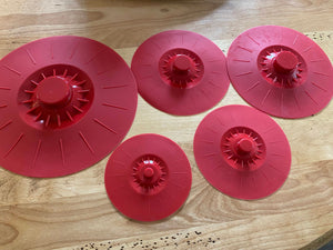 Five red silicone lids are laid out on a wooden table. The start with the largest, which could cover a large pot or bowl, and decrease in size.