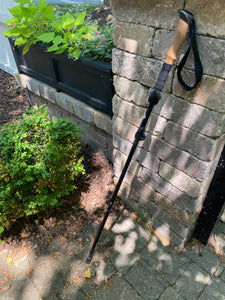 Black trekking pole with a cork handle and black wrist loop leaning against a brick wall
