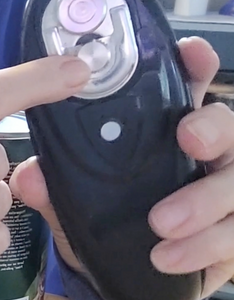 A person is pointing at the blade on the bottom of the can opener. The small magnet which helps lift the lid off after cutting is also visible.