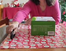 Load image into Gallery viewer, A person is seated at a table with a large piece of red and white wrapping paper over it, with a box on top. Their arm is stretched across the table using the cutter to cut across the entire piece of wrapping paper.
