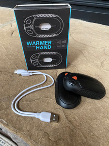 Two black hand warmers that look like computer mouses are sitting on a stone step. Beside them is a charging cord with two connectors so that they can be charged at the same time, as well as the box that they came in which says "Electric Warmer Hand"