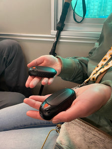 A person is holding out two black devices with an orange button on each one in the palms of their hands.