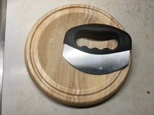 Load image into Gallery viewer, A mezzaluna (rocker) knife with a black, closed handle and a curved steel blade sitting on a circular wood cutting board that is slightly curved in the middle.
