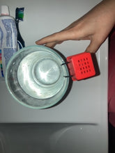 Load image into Gallery viewer, Close-up view of a glass with a metal rim at the top and a person&#39;s hand beside the glass. Attached to the rim is a red plastic object with two black wires protruding from it. The background includes a blurred view of a tube of toothpaste and a blue object, possibly a toothbrush or another type of tube. The focus is mainly on the red plastic object and the top of the glass container.
