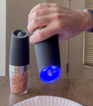 Load image into Gallery viewer, A person is tilting the pepper mill downward, activating the grinder and the blue light, as pepper comes out. The salt grinder, filled with coarse pink salt, is sitting on the table.
