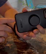 Load image into Gallery viewer, A person pointing at the charging port on the case. The brand &quot;otterbox&quot; is visibly printed on the bottom of the black case. The popsocket is also visible.
