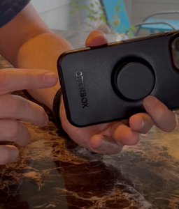 A person pointing at the charging port on the case. The brand "otterbox" is visibly printed on the bottom of the black case. The popsocket is also visible.