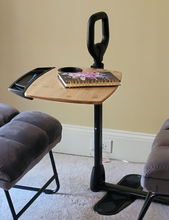 Load image into Gallery viewer, Bamboo tray table with a black tray attachment, a black cup holder, and a black stand assist handle positioned between a gray chair and a foot rest in a carpeted room.
