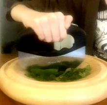 Load image into Gallery viewer, A person cutting herbs with a black mezzaluna knife with a steel blade on a wooden, curved cutting board.
