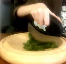 Load image into Gallery viewer, A person cutting herbs with a black mezzaluna knife with a steel blade on a wooden, curved cutting board.
