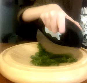 A person cutting herbs with a black mezzaluna knife with a steel blade on a wooden, curved cutting board.
