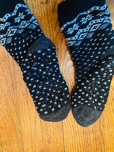 A pair of socks laying on a wood floor. There is a nice, winter design on them with white snowflakes and pattern at the top around the ankles and hten white dots over all over the foot. There are black grips that are not very visible.