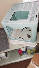Load image into Gallery viewer, The covered litter box is sitting on a shelf. The side entrance and the top entrance of the litter box are visible, as are the slats that cover the top of the box. The plastic door is completely open (the clear plastic door is not being used).
