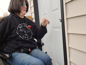 A woman with brown hair, sunglasses, and a black sweatshirt, is pulling the key toward the lock on the door, showing how the cord extends from the keychain attached to her purse. 