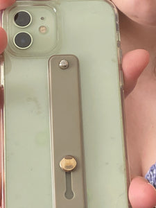 The olive green strap in the flat position, secured to the back of a phone. A silver button at the top is visible, and a slightly larger shiny button appears at the bottom, where space in the strap allows it to be pulled up or pushed back down to lay flat.