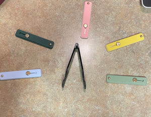 Five of the phone straps are laid out on the floor, all in different colors including light blue, dark green, pink, yellow, and light green. The free kickstand is black and sits in the middle.