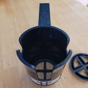 View of whole product, including the long thin black extension that slips under a seat cushion to keep the holder in place. The insert to raise smaller mugs to a higher level is sitting beside the cup holder on the table.