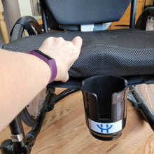 Load image into Gallery viewer, A person&#39;s hand is lifting the cushion to show that the HandiCup is held in place by a plastic extension that slips underneath the wheelchair seat cushion. 
