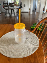 Load image into Gallery viewer, Plastic mason jar cup with handle sits on a placement on a table. The cup is clear with a neon orange lid and straw.

