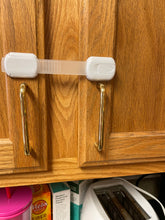 Load image into Gallery viewer, Two white pieces are each secured to a cabinet door, with a clear plastic piece stretching between them. Neither door can be opened without first detaching one end of the lock. 
