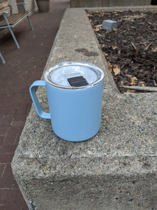 Blue mug with clear lid and black tab that opens the hole from which to drink. The mug is on a concrete planter wall.