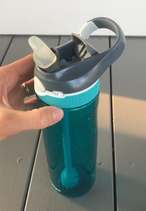 The bottle has a turquoise, see-through base with a gray lid with the spout open. The spout has a clear, thick cover over it. There is a fixed, plastic two-finger "loop" that is part of the lid. A white button is visible on the front, used to release the spout. A small slide button is visible, used to lock the spout. The bottle is sitting on the ground with a person's hand holding it.