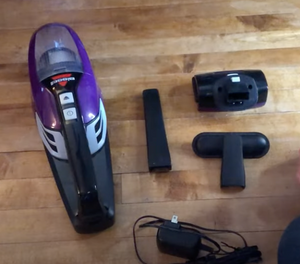 Purple and black Bissell Hand Vacuum on a wood floor. Beside it are three attachments and a charging cable, all black.