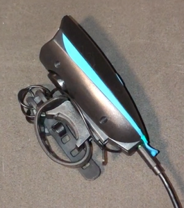 Side view of front light plugged into charging cable. Device is black with light blue accent and has a round loop strap with notches for attaching to device. 