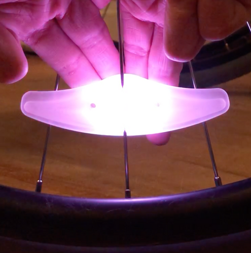 Spoke light is attached to wheel spokes and lit up pink. A person's fingers are pushing button on the back of the device.