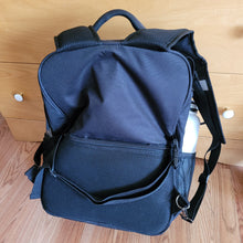 Load image into Gallery viewer, View of the back of the backpack with the straps lifted, revealing another strap that goes horizontally across the bag, as well as a &quot;hidden&quot; pocket, for which the zipper is half way up the back of the bag. 

