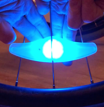 Load image into Gallery viewer, Spoke light lit up in blue. It is attached to the spokes of a wheel and a person is pushing a button on the back using several fingers.
