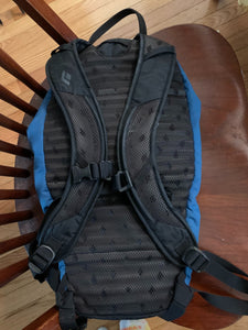 The back of the backpack, which is black with black straps and has a mesh material over the padding. There is a snap buckle visible at the middle of the straps and a handle on top of the bag. 