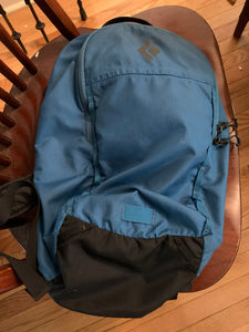 Blue backpack with a black bottom, black metal zipper pulls and a black diamond logo on the top front. One outside zipper is visible.