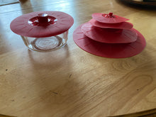 Load image into Gallery viewer, A red silicone lid with a visible popped handle in the middle on a clear food storage dish. Next to the dish are four lids of different sizes stacked on one another.
