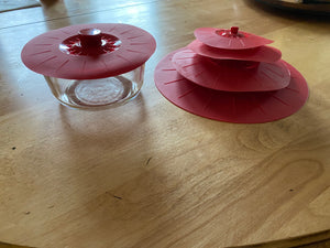 A red silicone lid with a visible popped handle in the middle on a clear food storage dish. Next to the dish are four lids of different sizes stacked on one another.