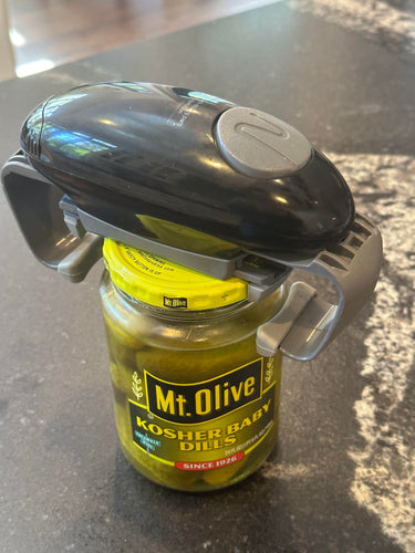 A black device with a large gray button and two gray pieces on the sides is sitting on top of a jar of Mt. Olive Kosher Baby Dill pickles on a countertop.