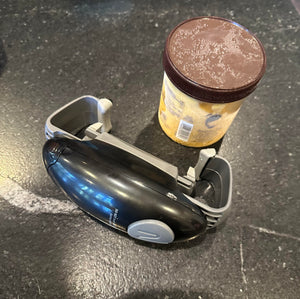 A black device with a gray button and gray pieces on its sides is on its side on a counter sitting beside a small tub of ice cream.