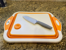 Load image into Gallery viewer, White cutting board with orange trim and drain plug sitting on a counter top. A knife with a gray handle and a plastic sheath is laying on the cutting board.
