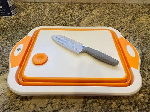 White cutting board with orange trim and drain plug sitting on a counter top. A knife with a gray handle and a plastic sheath is laying on the cutting board.