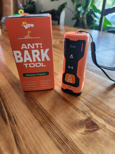 The Anti Bark Behavior Trainer Tool orange box is sitting on a table beside the device. The device has three buttons on the front, each with a different symbol. The two buttons are black and the top is orange. There is a black strap coming from one end of the device.