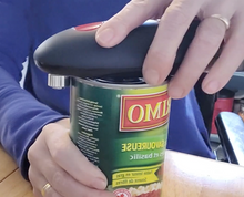 Load image into Gallery viewer, A person is placing the black can opener on a can of pasta sauce, with the metal piece resting on the outside of the can.
