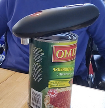 Load image into Gallery viewer, The can opener is operating on its own, with the metal piece on the outside of the rim of the can. The body of the device is slightly lifted up, which happens when it is working.
