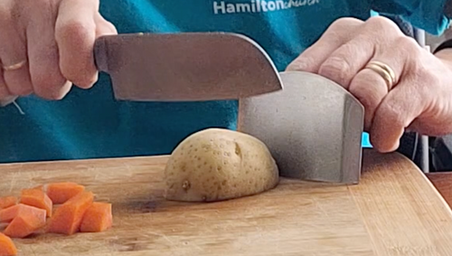 A person is using a small metal finger shield to steady a potato on a cutting board while using the other hand to chop the potato with a knife.