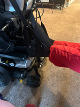 Load image into Gallery viewer, The black portable travel foot rest is hanging from a power chair. A person wearing black socks and red pants has their feet propped in the foot rest..
