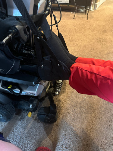 The black portable travel foot rest is hanging from a power chair. A person wearing black socks and red pants has their feet propped in the foot rest..