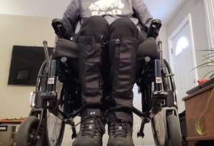 A person is seated in a wheelchair but we only see the lower half of their body. They are wearing black boots with black leg gaiters that go from the boot to below the knees.