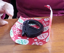 Load image into Gallery viewer, The black gift wrap cutter is on its side on top of a small piece of wrapping paper. It has a relatively large handle and the blade is not visible.
