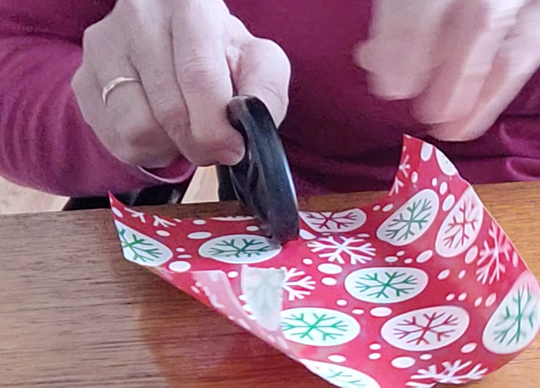 A person is using a black gift wrap cutter to cut a piece of red gift paper with snowflakes.