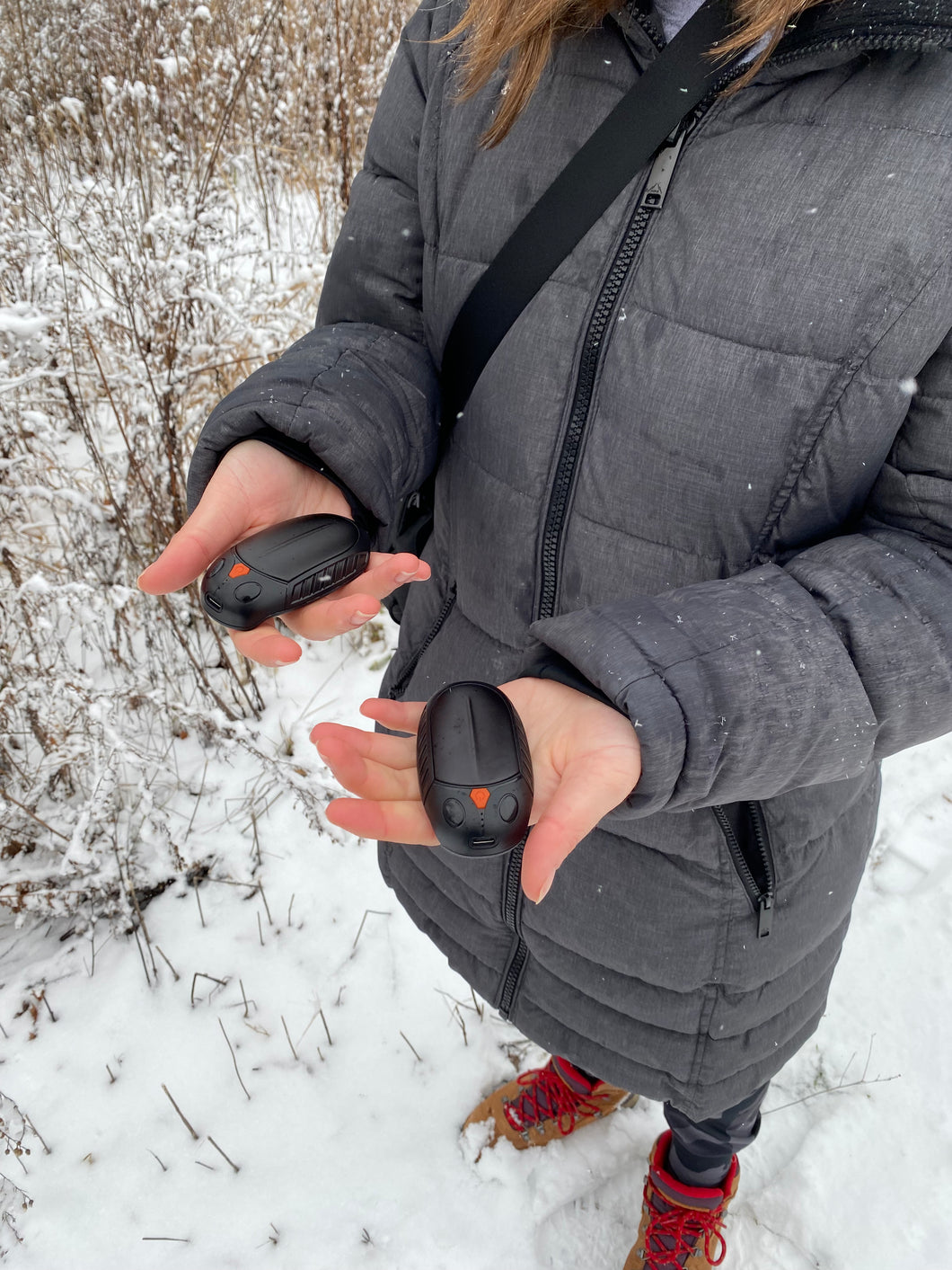 A person is holding two round, black devices with an orange button in the middle of each. One is in each hand. There is snow all around.